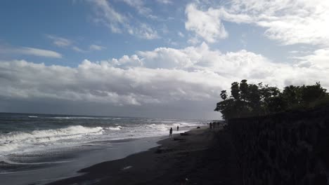 Black-Sand-Cliff-and-Sea-Water-at-Saba-Beach-Blahbatuh,-Gianyar-Bali-Indonesia,-People-with-their-feet-on-the-Ocean-Shore,-Afternoon-Time,-Blue-Sky