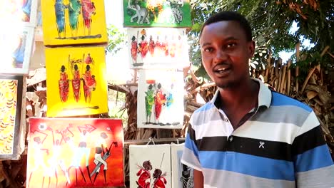 African-vendor-explaining-to-the-camera-Tingatinga-drawings-on-display-for-sale-to-tourists-in-Arusha,-Tanzania