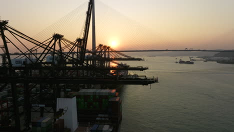 Gantry-cranes-loading-and-discharging-containers-on-moored-ships-with-a-large-suspension-bridge-in-background-during-sunset
