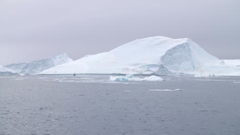 Slow-motion-pan-across-icebergs,-ice-floes,-and-a-fishing-boat-on-the-ocean-beneath-a-cloudy-sky