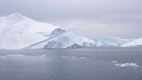 Slow-motion-pan-of-a-large-iceberg-with-small-floes-of-ice-in-front-of-it---floating-on-a-calm-ocean-under-a-cloudy-sky-off-the-coast-of-Greenland