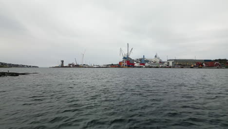 Low-drone-over-sea-toward-Rosenberg-shipyard-with-cranes,-Stavanger,-Norway