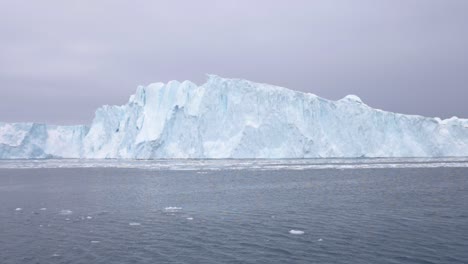 Cámara-Lenta-De-Un-Gran-Iceberg-Flotando-En-El-Océano,-Con-Pequeños-Témpanos-De-Hielo-En-Primer-Plano-Bajo-Un-Cielo-Nublado-Frente-A-La-Costa-De-Groenlandia