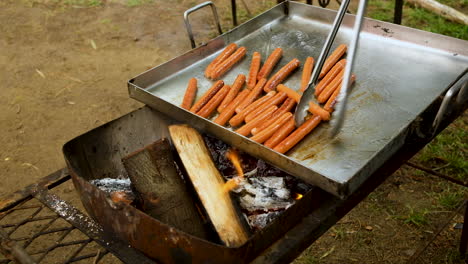 Frying-sausages-on-big-camping-pan-on-open-wood-fire-using-tongs