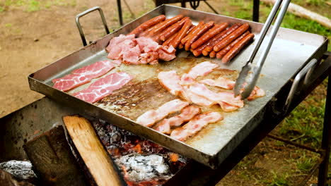 Frying-bacon-and-sausages-on-stainless-steel-pan-on-open-fire---camp-breakfast