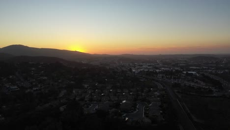Drone-captures-a-breathtaking-aerial-shot-of-the-sun-setting-on-the-small-town,-rising-higher-and-higher-as-the-colors-of-the-sky-change-and-the-setting-light-reaches-over-the-horizon