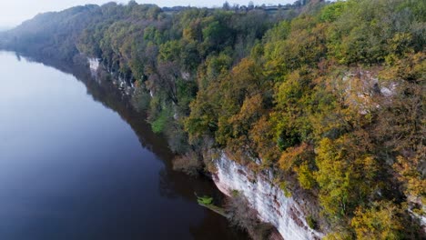FPV-Drone-flying-along-a-cliff-over-a-river-with-trees-in-autumn,-Dordogne-River---Bac-de-Sors-in-France