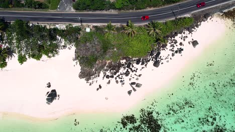 Mahe-Seychelles-drone-shot-of-car-passing-on-the-road-near-the-beach,-palm-tees-and-granite-rocks-can-be-seen,-white-sandy-beach-and-turquoise-water