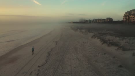 Aerial,-man-running-on-the-beach-early-in-the-morning,-sea-mist-rolling-inland