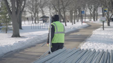 On-a-bright-winter-day-in-a-park,-a-volunteer-wearing-a-high-visibility-vest-carries-a-crowd-control-barrier,-slow-motion