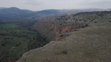 Aerial-drone-flyover-rocky-terrain-revealing-green-valley-below,-Israel,-Nahal-Amud