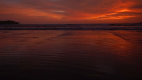 Panoramablick-Auf-Die-Seelandschaft-Mit-Meereswellen,-Die-An-Der-Sandigen-Küste-Zusammenbrechen,-Während-Eines-Leuchtend-Roten-Und-Orangefarbenen-Sonnenuntergangs-An-Einem-Bewölkten-Abend