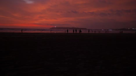 Blick-Auf-Das-Meer-Von-Menschen-Silhouetten-An-Einer-Sandigen-Küste,-Während-Eines-Leuchtend-Roten-Sonnenuntergangs,-An-Einem-Bewölkten-Abend