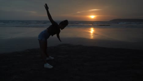 Slow-motion-landscape-view-of-a-young-woman-silhouette-doing-stretching-exercises,-on-a-sandy-beach-by-the-ocean,-at-sunrise