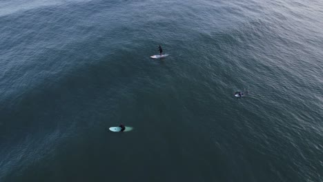 Drone-flying-above-the-sea,-Two-surfers-waiting-for-waves-and-a-standup-paddle