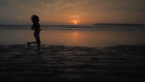 Landscape-view-of-a-silhouette-of-a-young-woman-running-on-a-sandy-beach-by-the-ocean,-at-sunrise