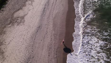 Drone-flying-over-and-behind-a-surfer-walking-in-the-shore-with-his-board-behind-his-arm