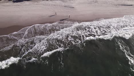 Static-drone-shows-the-shore-when-two-men-passes-each-other-near-the-waves,-one-with-his-surf-board-over-his-head