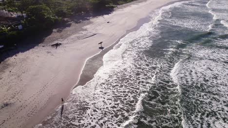 Drone-flying-over-the-shore,-couple-of-surfers-walking-near-the-waves-with-their-boards