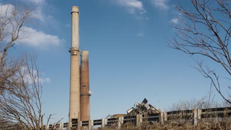 Old-coal-fired-power-plant-smoke-stacks-against-a-blue-sky-with-trees-and-road-in-foreground,-demolished-building