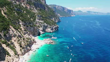 Cala-Mariolu-Beach,-Tourists-and-Boats-at-Baunei-Coast,-Sardinia,-Italy---Aerial-4k