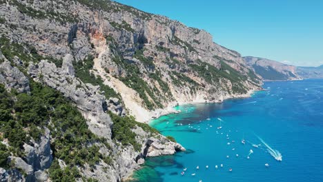 Baunei-Coast,-Cala-Goloritze-Beach-and-Tourist-Boats-at-Turquoise-Blue-Sea-in-Sardinia,-Italy---Aerial-4k