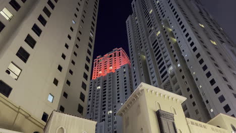 Wide-view-from-the-ground-of-a-tall-tower-with-red-lights-at-the-top-in-Dubai-city-at-night