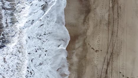 Cenital-spiral-drone-shot-of-the-sea-waves-breaking-on-the-beach-with-tracks-on-the-sand,-in-Cariló,-Argentina