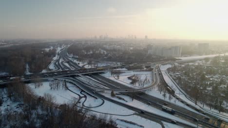 Established-aerial-footage-moving-forward-of-a-traffic-intersection-road-covered-in-snow-with-the-skyline-of-the-city-center-of-Warsaw-at-sunset-in-winter