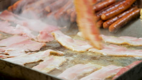 Close-up-of-flipping-bacon-with-tongs-that's-frying-on-pan,-breakfast-prep