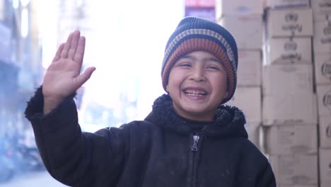 Young-Male-Kid-Wearing-Hat-Smiling-And-Waving-With-Hand-Looking-Directly-At-Camera-In-Street-In-Quetta,-Balochistan
