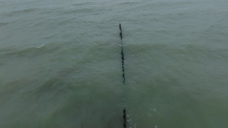 Aerial-birdseye-view-of-an-old-wooden-pier-at-the-Baltic-sea-coastline,-overcast-winter-day,-white-sand-beach,-wooden-poles,-waves-hitting-shore,-wide-drone-shot-moving-backward-low