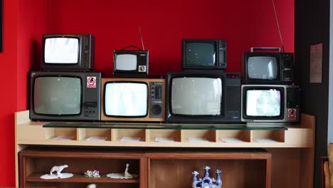 Montones-De-Televisores-Antiguos-Con-Pocos-Encendidos-En-Un-Estante-De-Madera-Contra-La-Pared-Roja
