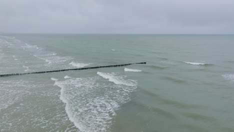 Aerial-establishing-view-of-an-old-wooden-pier-at-the-Baltic-sea-coastline,-overcast-winter-day,-white-sand-beach,-wooden-poles,-waves-hitting-shore,-wide-drone-shot-moving-forward