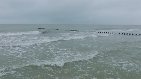 Aerial-establishing-view-of-an-old-wooden-pier-at-the-Baltic-sea-coastline,-overcast-winter-day,-white-sand-beach,-wooden-poles,-waves-hitting-shore,-wide-drone-shot-moving-forward-low