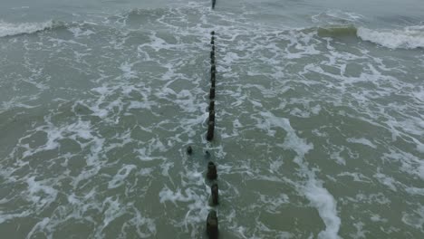 Aerial-birdseye-view-of-an-old-wooden-pier-at-the-Baltic-sea-coastline,-overcast-winter-day,-white-sand-beach,-wooden-poles,-waves-hitting-shore,-wide-drone-shot-moving-forward-low