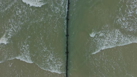 Aerial-birdseye-view-of-an-old-wooden-pier-at-the-Baltic-sea-coastline,-overcast-winter-day,-white-sand-beach,-wooden-poles,-waves-hitting-shore,-wide-drone-shot-moving-forward