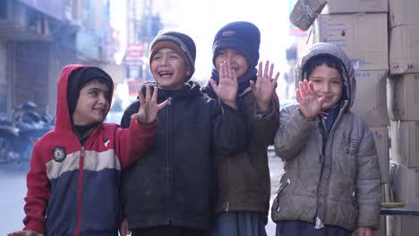 Group-Of-Young-Male-Kids-Wearing-Hat-Smiling-And-Waving-With-Hand-Looking-Directly-At-Camera-In-Street-In-Quetta,-Balochistan