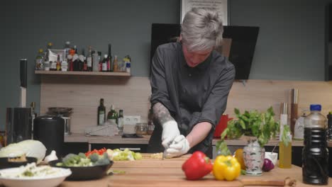 Young-professional-male-chef-in-an-elegant-black-shirt-with-an-alternative-look,-tattoos,-gloves-slicing-fresh-leek-A-modern,-fancy-looking-kitchen-with-fresh-vegetables-besides-Cinematic-Dolly-4K