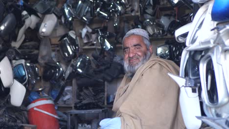 Älterer-Ladenbesitzer,-Der-In-Einem-Autohaus-Für-Ersatzwagen-In-Quetta,-Pakistan,-Sitzt-Und-In-Die-Kamera-Lächelt