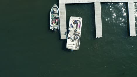 Aerial-top-down,-pontoon-boat-leaving-docking-pier-on-a-lake-during-the-day