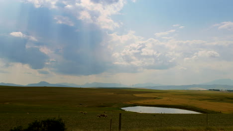 Panorama-Bauernhof-Landschaftsaufnahme---Schafe-Grasen-In-Der-Nähe-Eines-Kleinen-Damms-Mit-Sonnenstrahlen,-Die-Durch-Wolken-Auf-Windturbinen-Brechen