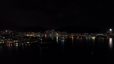 Un-Barco-Sale-Del-Puerto-De-Bergen-Mientras-Se-Ven-Fuegos-Artificiales-Por-Toda-La-Ciudad-Durante-La-Celebración-Del-Año-Nuevo---Antena-Panorámica-Nocturna---Noruega
