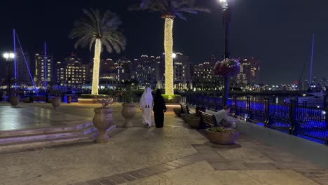 Arabisches-Paar-Mann-Frau-Mit-Einem-Traditionellen-Lokalen-Modekleid-Bisht-Hijab-Wandernder-Luxusstrand-In-Katara-Perle-Qanat-Quartier-In-Doha-Katar-Das-Stadtlandschaftsdesign-Wird-Von-Palmen-Und-Licht-Geschmückt