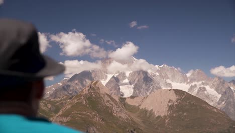 Mann-Mit-Hut,-Der-An-Einem-Sonnigen-Tag-In-Den-Alpen-Auf-Einen-Großen-Berggipfel-Blickt,-Der-Mit-Wolken-Bedeckt-Ist