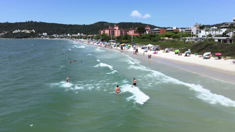 Aerial-scene-of-drone-flying-low-over-the-sea-and-waves-with-many-people-diving-and-having-fun-on-the-beach-sand-with-many-buildings-and-hotels-facing-the-sea-in-Florianópolis