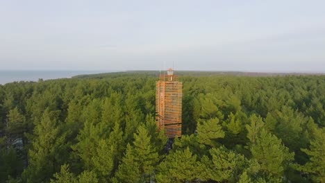 Aerial-view-of-Bernati-lighthouse-surrounded-by-lush-green-pine-tree-forest-with-light-snow,-Nordic-woodland,-Baltic-sea-coast,-sunny-winter-day,-Latvia,-wide-ascending-drone-shot-moving-backward