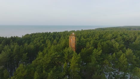 Aerial-view-of-Bernati-lighthouse-surrounded-by-lush-green-pine-tree-forest-with-light-snow,-Nordic-woodland,-Baltic-sea-coast,-sunny-winter-day,-Latvia,-wide-drone-shot-moving-forward,-tilt-down