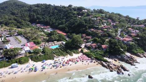 Aerial-drone-scene-of-residential-neighborhood-facing-the-beach-with-lots-of-nature-sand-sea-and-rocky-shores-with-many-people-having-fun-on-the-beach-many-umbrellas-in-summer-florianopolis-jurere