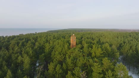 Aerial-view-of-Bernati-lighthouse-surrounded-by-lush-green-pine-tree-forest-with-light-snow,-Nordic-woodland,-Baltic-sea-coast,-sunny-winter-day,-Latvia,-distant-wide-orbiting-drone-shot